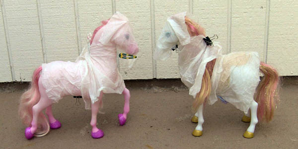This photo shows a side view of two toy horses facing each other. Each horse has its head, chest, and back draped by damp paper towels that are clipped with hair clips or binding clips.