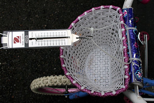 A spring scale is attached to the center of a bikes handlebars using plastic zip ties