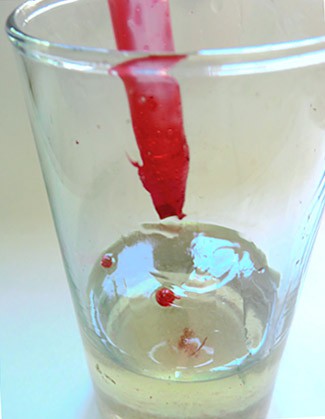 Red gelatin in a syringe is dropped into a cup filled with oil