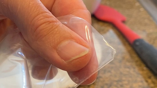 A bottom corner of a resealable plastic bag with a small tip cut off.  The  cut-off tip is about 2 millimeters wide.   