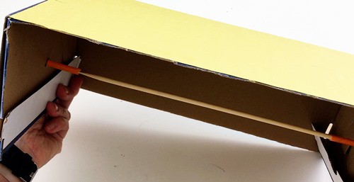 a skewer treaded through two short pieces of a straw sticking out from opposite side panels of a shoebox. 