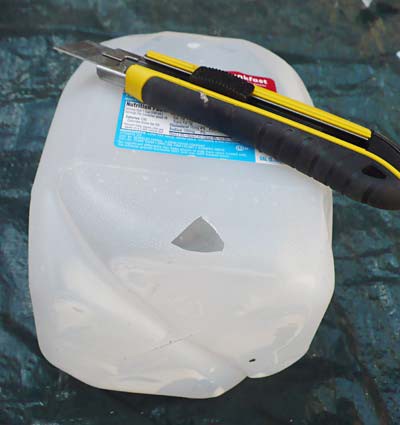 A utility knife is used to cut a triangular hole in the bottom of a plastic water jug