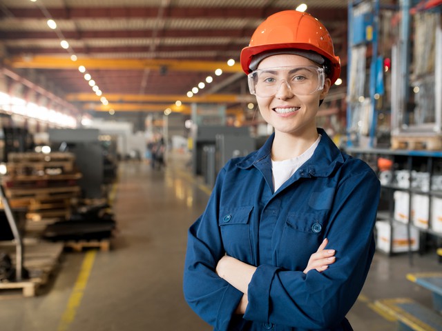 Young smiling female technician in blue uniform, protective eyeglasses and helmet working in modern factory