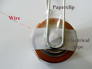 Wire is taped to the positive terminal of a battery and a paperclip is taped over the tape holding the wire