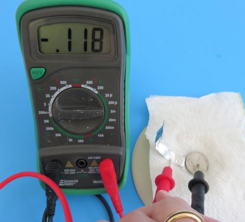 A multimeter measures the voltage in milliamps across an aluminum foil strip and a stack of a penny, paper towel, and nickel