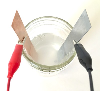 Alligator clips attach to a copper strip and zinc strip which both rest in a glass filled with saltwater