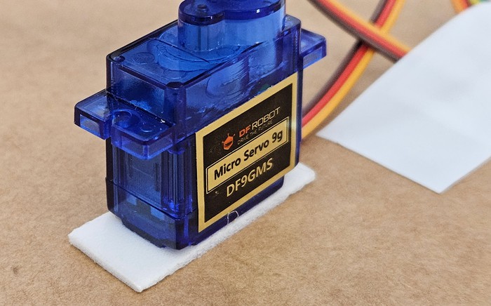 Micro servo motor with sitting upright on a piece of double-sided foam tape which is on top of a piece of cardboard 