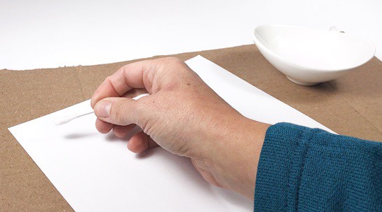 A hand holding a Q-tip and writing a message onto a white sheet of paper.