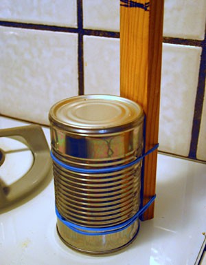 An upside-down steel can is attached to a wooden stick with rubber bands