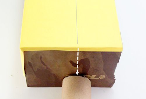 A cardboard tube pressed against the side panel of a shoebox so that the bottom of the tube aligns with the lower edge of the box. A vertical arrow extends from line on top of the box to the top of the tube.