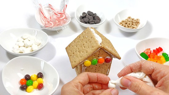 A Graham cracker house is standing between small bowls filled with assorted candies. 