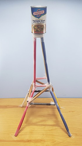  A paper tower with a truss structure that is wider at the bottom and narrow at the top, with a can of beans at the top. 