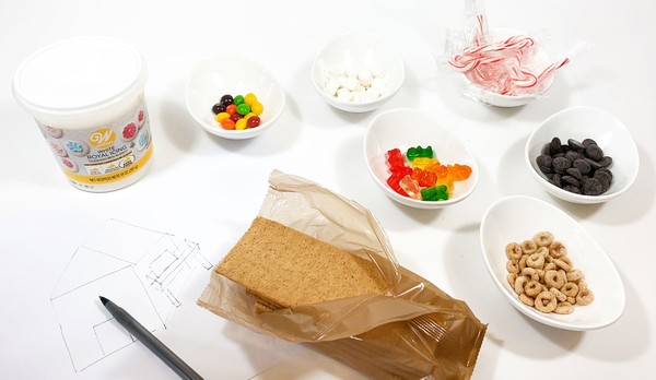  Materials needed for the 'Gingerbread House Engineering Challenge' activity: Royal icing, Graham crackers, a paper, pencil, and and assortment of candies on a table.