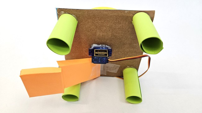 Square piece of cardboard with four cylindrical legs and a servo motor glued to the center of the cardboard