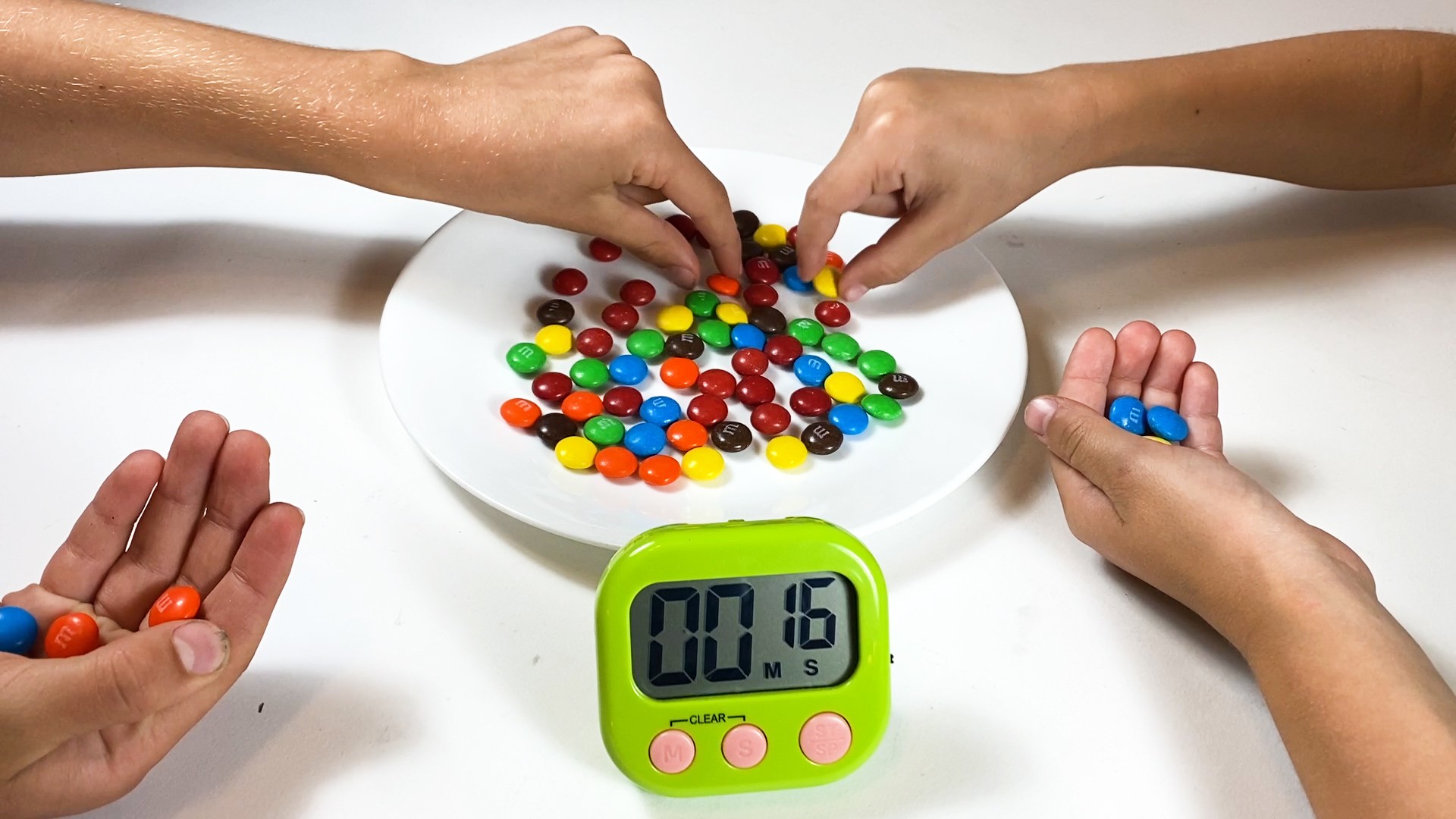 Multiple hands pick up M&M candies off of a plate with a timer set
