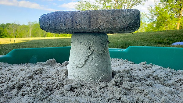 A bucket-shaped sandcastle in a sandbox with some pieces of grass sticking out and a brick resting on top. 