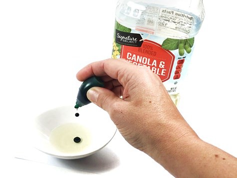 Hand squeezing food color from a bottle into a small bowl filled with cooking oil.