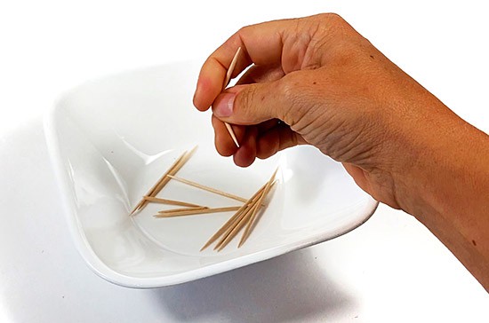 A hand holding a toothpick above a bowl filled with toothpicks.