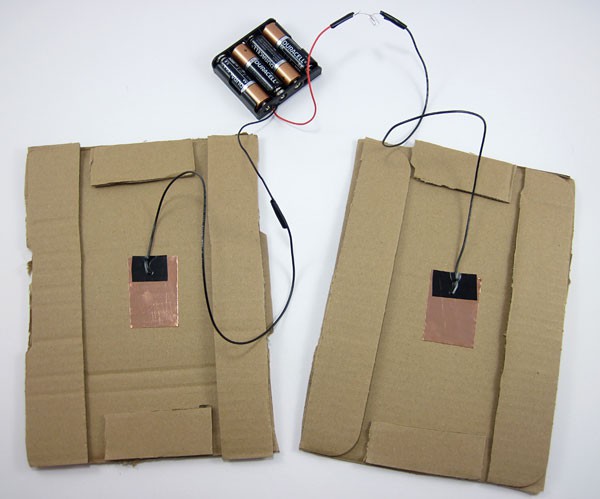 A battery pack connects to two copper plates which are taped to the inside of separate cardboard sheets