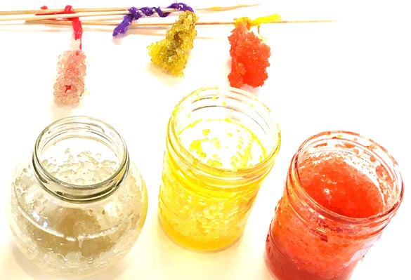 Three jars with large sugar crystals sticking to the inside of the jars, and three strings with rock candy attached to them. Each rock candy is a collection of large sugar crystals sticking to each other and to the string. They look similar to rock-formations found in nature.  