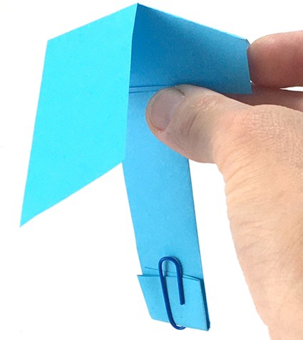 Person holding the paper helicopter in the middle with the triple folded part hanging vertically down.  