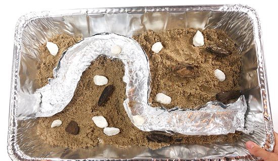 Aluminum pan with curved aluminum riverbed inside surrounded by sand, pebbles and wood chips.