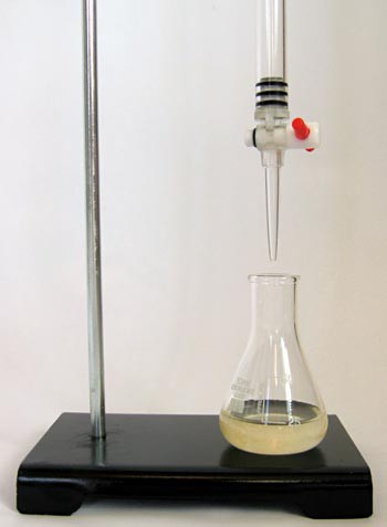 An erlenmeyer flask filled with a light orange solution is placed under a buret