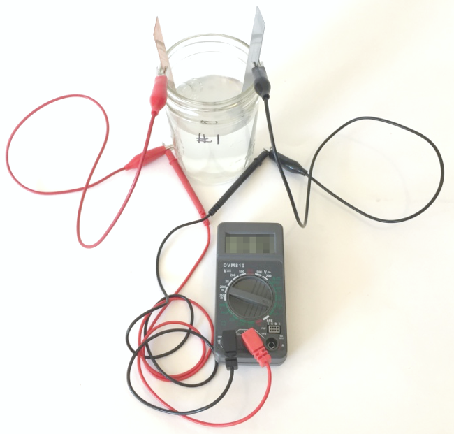 Alligator clips attach multimeter probes to a copper strip and zinc strip which both rest in a glass filled with saltwater