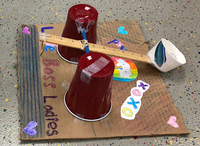 A student next to a ball launcher made from plastic cups, pencils, a ruler and tape