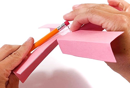 Hands holding a paper pinwheel made from 4 paper strips. The paper pinwheel is attached to the pencil eraser with a thumbtack.  