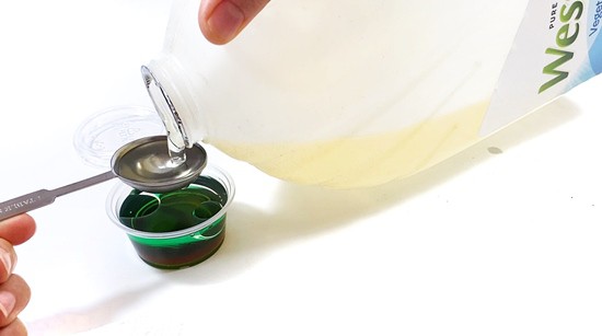 A mini cup with dark corn syrup and green water in it is standing on a table. A tablespoon is held above the mini cup and is filled with vegetable oil from a container.