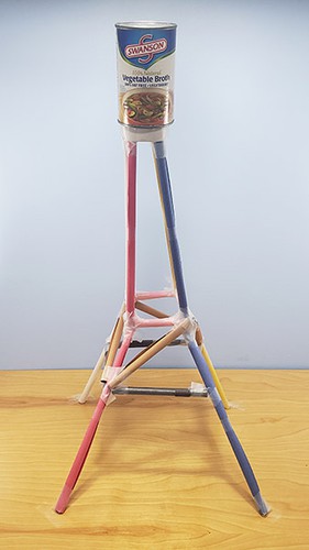 A paper tower with a truss structure that is wider at the bottom and narrow at the top, with a can of beans at the top.