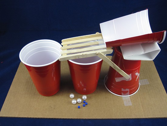 Different sized marbles next to popsicle sticks and a cut in half plastic cup that form a ramp over two plastic cups