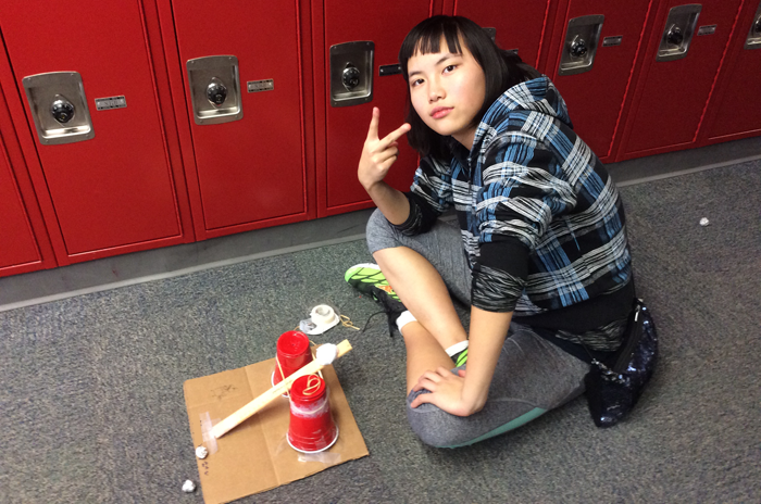 A student next to a ball launcher made from plastic cups, pencils, a ruler and tape