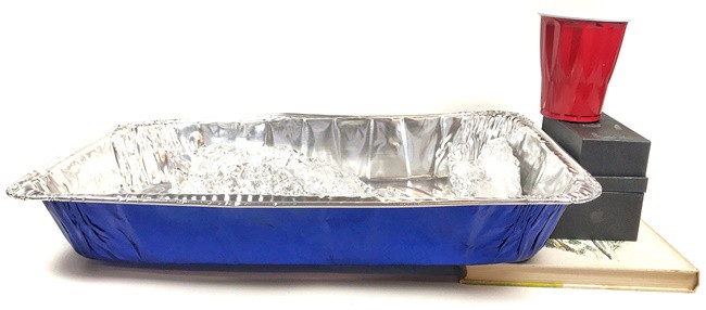 Inclined aluminum pan with the river model inside. A plastic cup is placed on top of boxes next to the aluminum pan, so that the water can flow from the cup into the aluminum river.