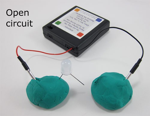 Two leads of a battery pack are inserted into two separate balls of Play-Doh, one of which has an LED partially inserted