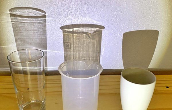 Image showing a glass, a frosted plastic beaker, and a ceramic cup standing in front of a wall. Light that shines on the objects projects shadows of each object on the wall. The glass shadow is very light, the frosted plastic beaker shadow is darker, and the shadow of the ceramic cup is black.