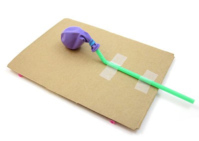 A straw secured to the opening of a balloon is taped to a sheet of cardboard