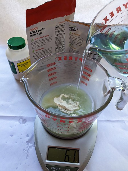 A pyrex measuring cup containing agar powder sitting on a scale while boiling water is poured into it to make a hydrogel.  