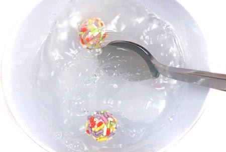 A bowl of soapy water is stirred with a spoon. Two different aluminum balls covered with sprinkles are floating on top of the water.