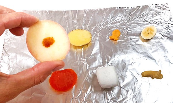 A variety of food pieces are spread on a sheet of aluminum foil. A hand is holding a piece of apple that has a drop of iodine solution on it.