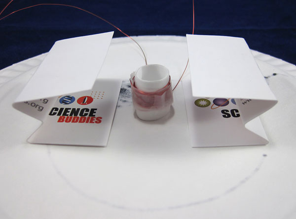 Two business cards bent into the shape of a sideways W sit on either side of a paper tube wrapped in copper wire coils