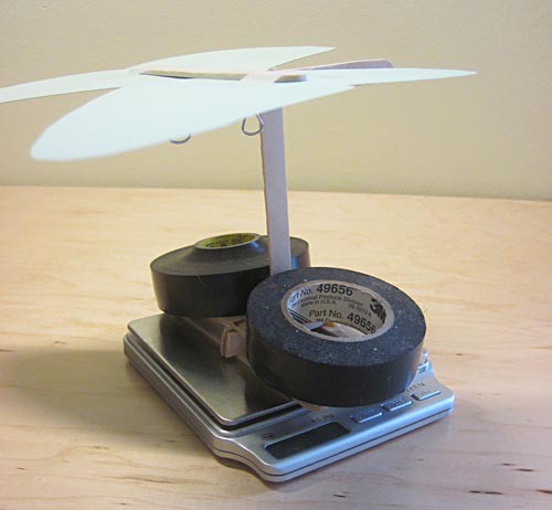 A paper butterfly cutout on a popsicle stick support structure and two rolls of electrical tape are placed on a scale