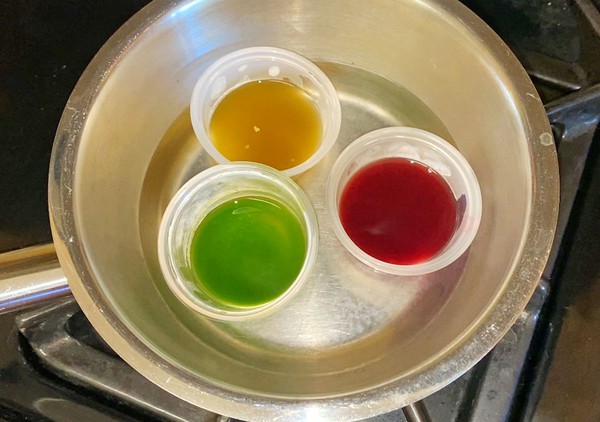 Three small cups are sitting in a pot filled with some water. Each cup contains a different colored leaf extract solution: green, yellow, and red.