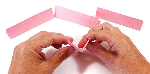 Three folded paper strips on a table. Hands holding one paper strip opening the fold so it looks like a V-shape. 