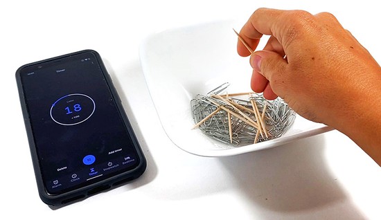  A hand holding a toothpick above a bowl filled with paper clips and toothpicks. A timer next to the bowl shows 18 seconds. 