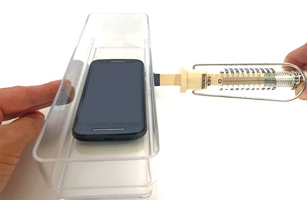 A spring scale pushing against a plastic container with a smartphone inside
