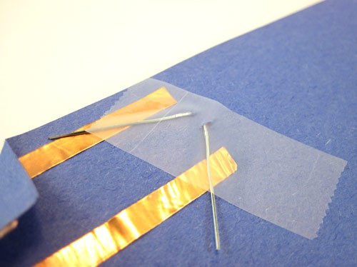 Two leads from an LED poke through a sheet of paper and are taped down onto two strips of copper tape