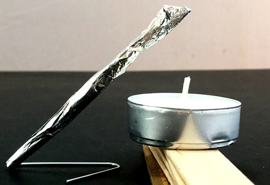 Rocket ready to launch from a paperclip launcher. A candle is resting on a stack of craft sticks so the flame would reach the tip of the rocket. 