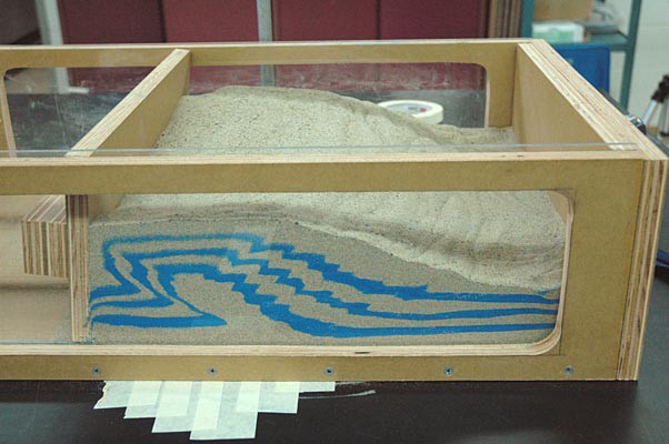 As a wood panel moves to compress sand, layers of blue sand are curved and bent